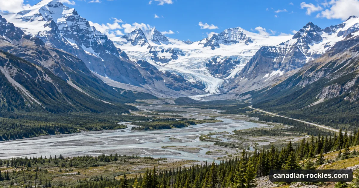 A wide glaciated valley with a braided river and a distant empty highway below snow peaks